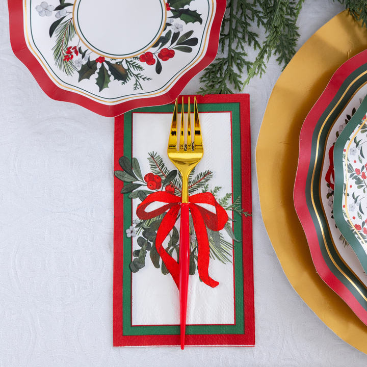 A festive Home for the Holidays: Christmas Table for 16 setting with holly-patterned heavy-duty paper plates, gold-accented disposable plates, and a red-handled gold fork on a mistletoe-decorated napkin with a red ribbon.