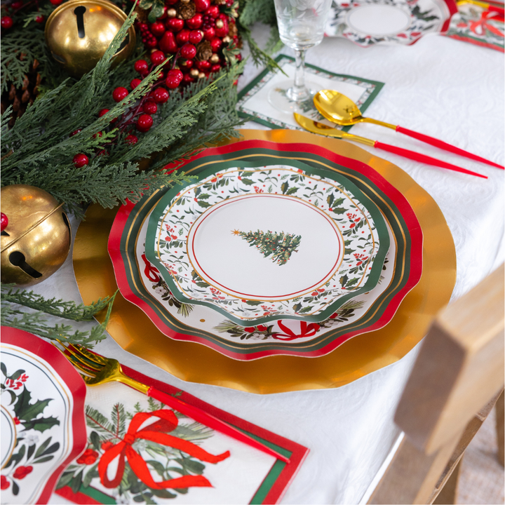 A Home for the Holidays: Christmas Table for 16 setting featuring tiered paper plates with tree and holly patterns, gold chargers, gold flatware with red handles, and a centerpiece of evergreen, berries, and large gold bells.
