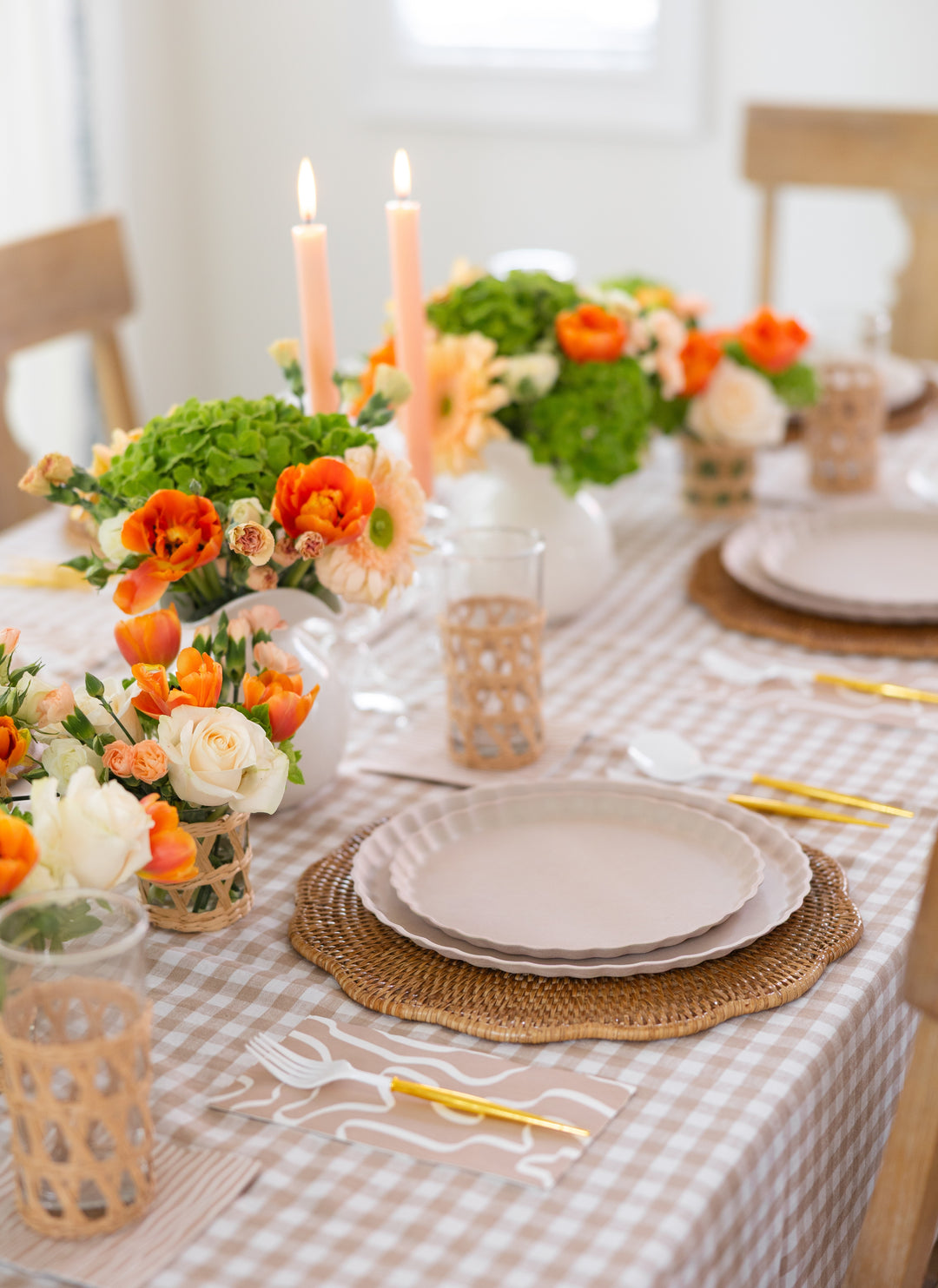 A dining table set for a meal with a gingham tablecloth, plates on woven placemats, gold cutlery, glasses with wicker holders, lit candles, floral orange and white centerpieces, and Sophistiplate Guest Towel Eco Sand napkins for an eco-friendly touch.