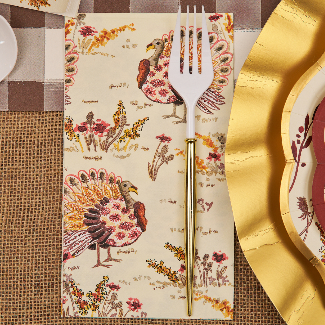 A gold and white fork rests on a Sophistiplate Amber Harvest Guest Towel, decorated with colorful turkeys and flowers, placed on a fall-themed table setting with gold-edged plates and a burlap and plaid tablecloth.