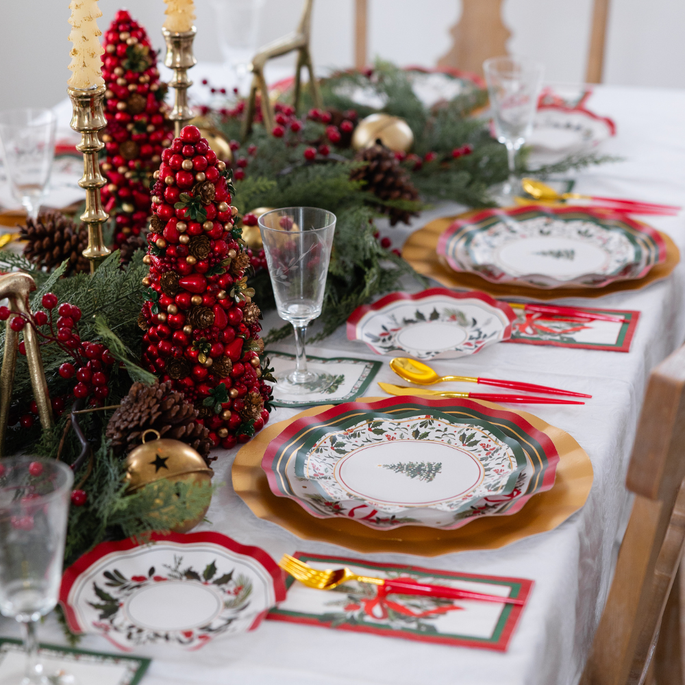 A Home for the Holidays: Christmas Table for 16 setting featuring layered heavy duty paper plates with evergreen patterns, gold-toned cutlery, red berry centerpieces, and decorative mini trees.