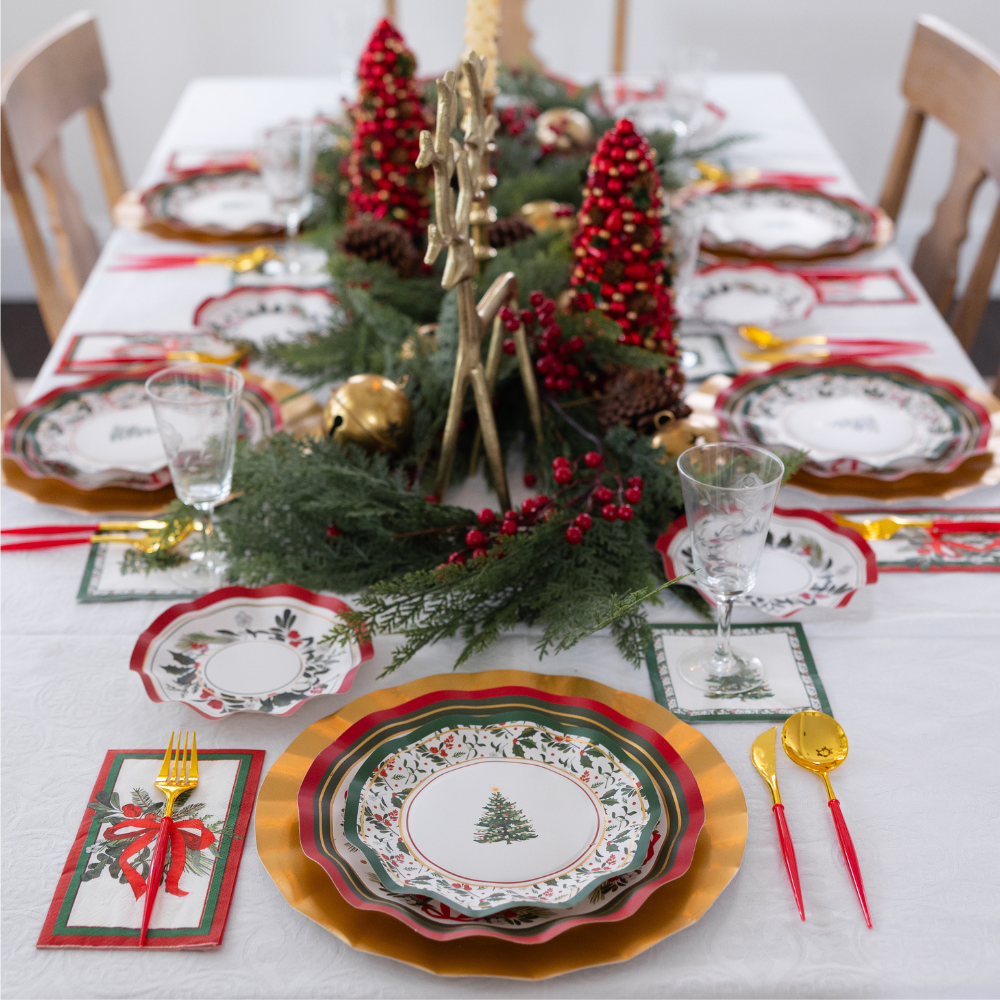 A Home for the Holidays: Christmas Table for 16 set features heavy-duty paper plates, gold-rimmed chargers, red-handled cutlery, and a centerpiece of evergreen branches, red berries, and miniature trees.