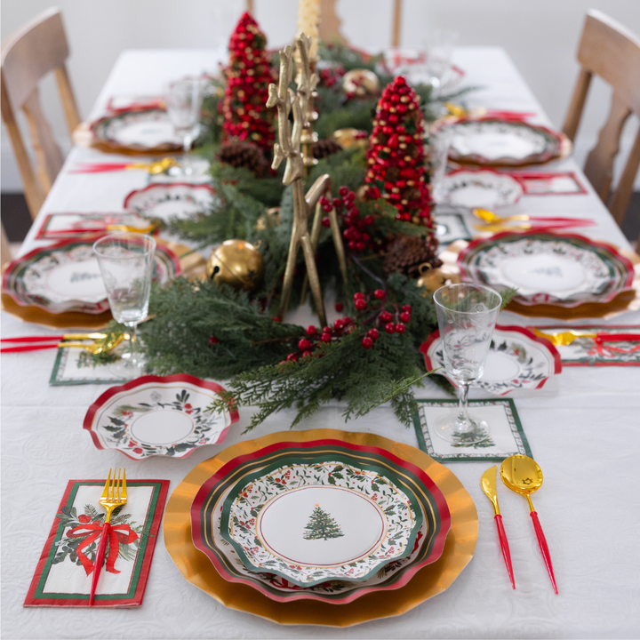 A Home for the Holidays: Christmas Table for 16 set features heavy-duty paper plates, gold-rimmed chargers, red-handled cutlery, and a centerpiece of evergreen branches, red berries, and miniature trees.