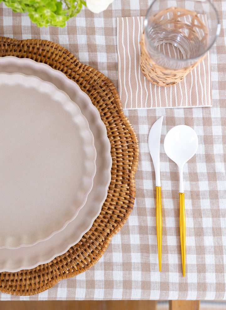 A table setting with scalloped ivory plates on a round wicker placemat, a glass in a woven holder, white flatware with gold handles, Sophistiplate Cocktail Napkin Eco Sand/20 pkg, all on a beige and white gingham tablecloth.