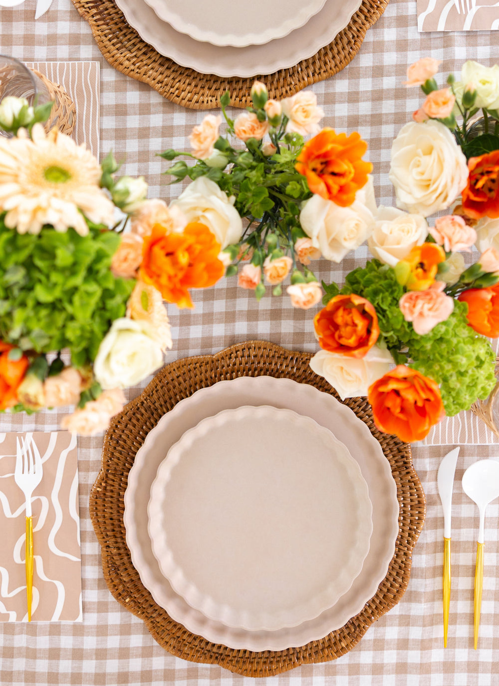 A top-down view of a table with Sophistiplate's Simply Eco Compostable Table Setting - Sand, featuring scalloped-edge biodegradable bamboo plates on woven placemats, gold utensils, and orange and white floral centerpieces on a beige gingham tablecloth.