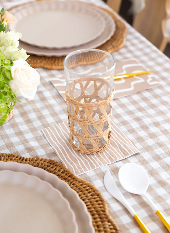 A close-up of a table set with a glass in a wicker holder, Sophistiplate Cocktail Napkin Eco Sand, white plate on a woven placemat, gold and white cutlery, and white flowers atop a beige checkered tablecloth.