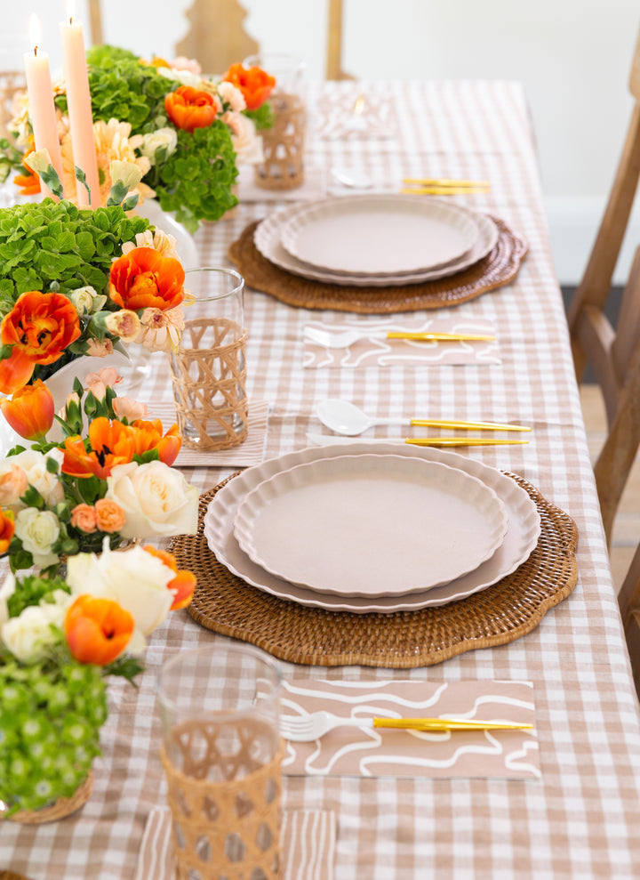 Sophistiplate’s Eco-Friendly Table Setting- Sand features a beige checkered tablecloth, biodegradable bamboo plates on woven placemats, gold cutlery, wicker holder glasses, and orange, peach, and green floral accents.