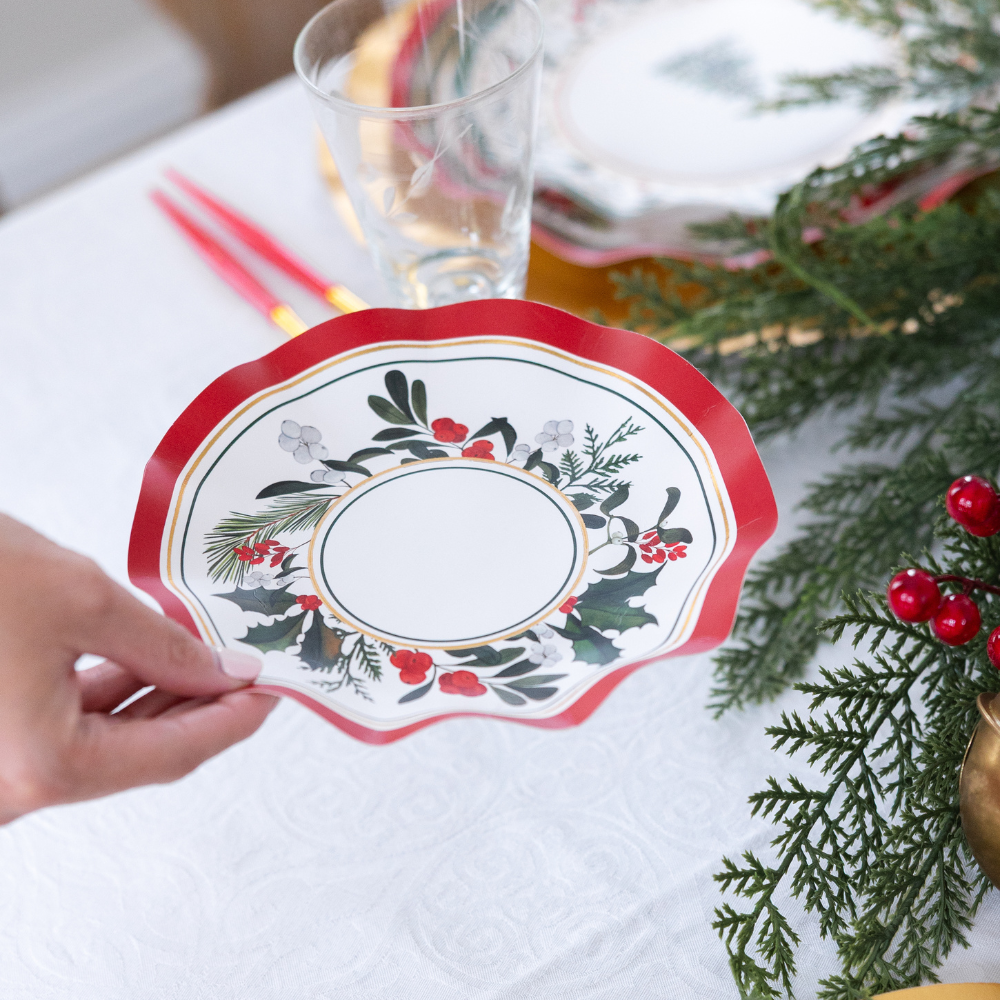 A hand holds a scalloped, red-rimmed Home for the Holidays: Christmas Table for 16 paper plate, decorated with a pattern of holly, berries, and pine sprigs.