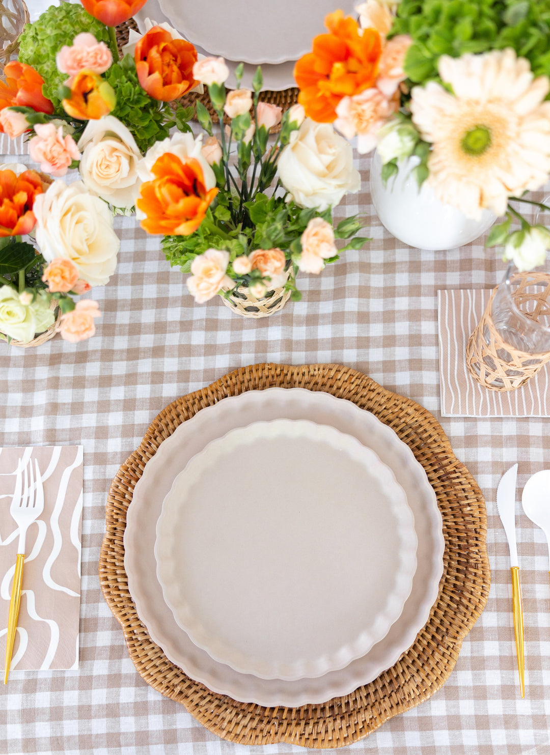 A table setting with stacked white plates on a woven placemat, gold cutlery, Sophistiplate Cocktail Napkin Eco Sand paper napkins, and vases of orange and white flowers atop a beige gingham tablecloth.