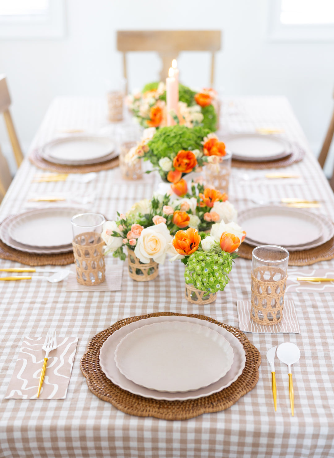 The Sophistiplate Eco-Friendly Table Setting- Sand features compostable plates, woven placemats, gold and white cutlery, patterned glasses, and floral accents on a beige gingham tablecloth with orange and white flowers.