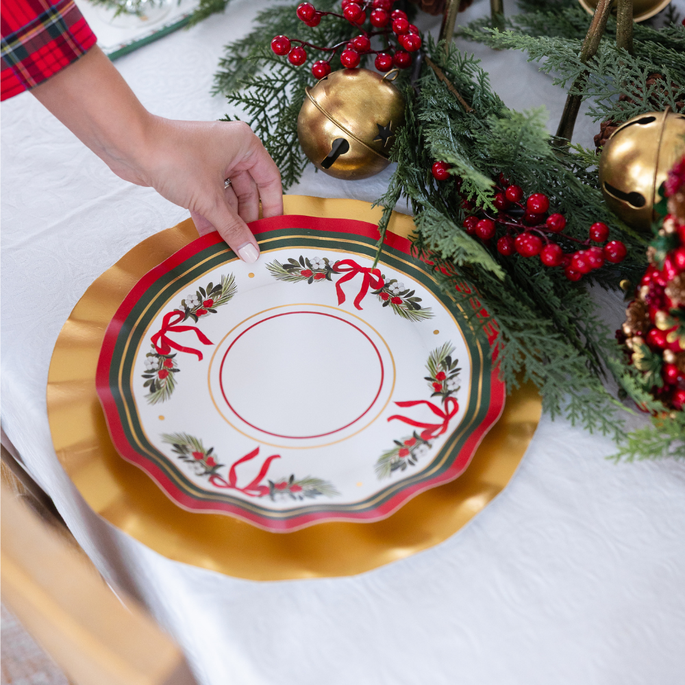 A hand places a Sophistiplate LLC Home for the Holidays: Christmas Table for 16 paper plate with a red ribbon and evergreen design onto a gold charger at a table decorated with pine branches, red berries, and large gold bells.