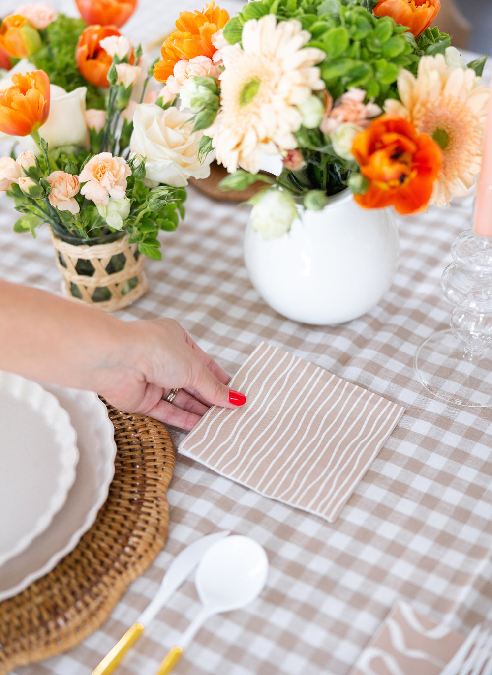 A hand with red nail polish sets Sophistiplate Cocktail Napkin Eco Sand/20 pkg on a gingham tablecloth, accented by white plates, gold utensils, and vases of orange, cream, and green flowers.