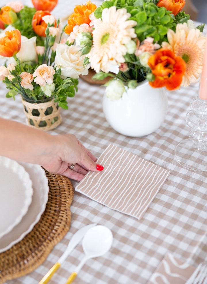 A hand with red nail polish sets Sophistiplate Cocktail Napkin Eco Sand/20 pkg on a gingham tablecloth, accented by white plates, gold utensils, and vases of orange, cream, and green flowers.