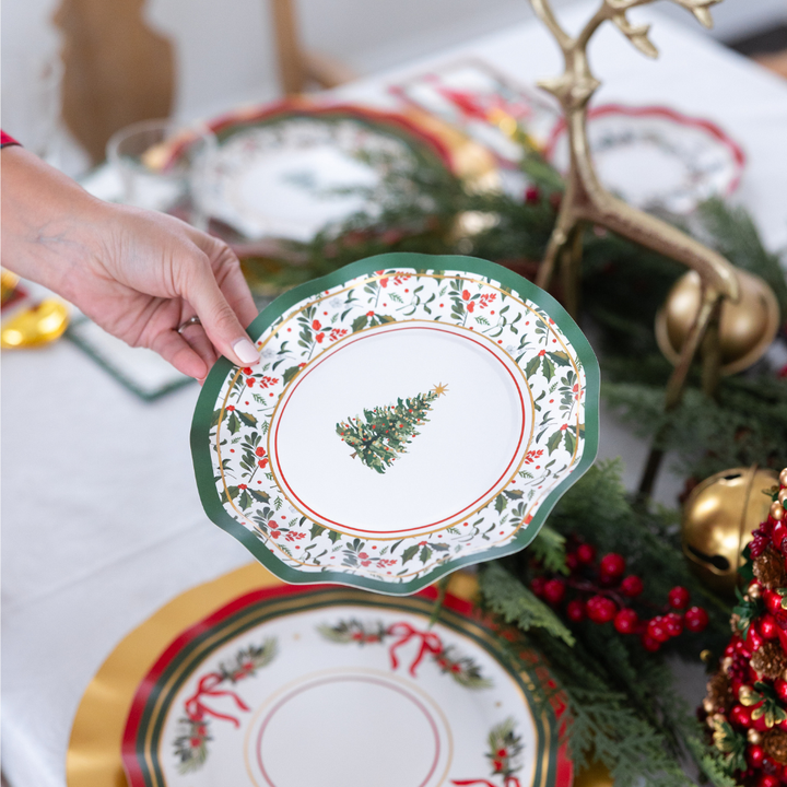 A hand holds a scalloped-edge Home for the Holidays: Christmas Table for 16 heavy-duty paper plate with a Christmas tree pattern above a festively set holiday table.