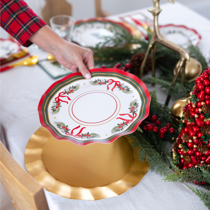 A hand places a Home for the Holidays: Christmas Table for 16 heavy duty paper plate, decorated with a red and green Christmas ribbon and holly pattern, onto a gold charger plate.