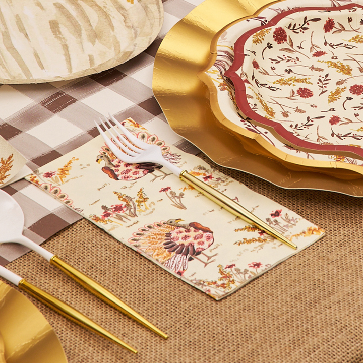 Thank you for your request. Here is the alt text, under 250 characters: Elegant fall table setting with Sophistiplate Amber Harvest Guest Towels featuring turkeys and autumn foliage, gold-edged plates, and gold-handled utensils on a checkered burlap tablecloth.