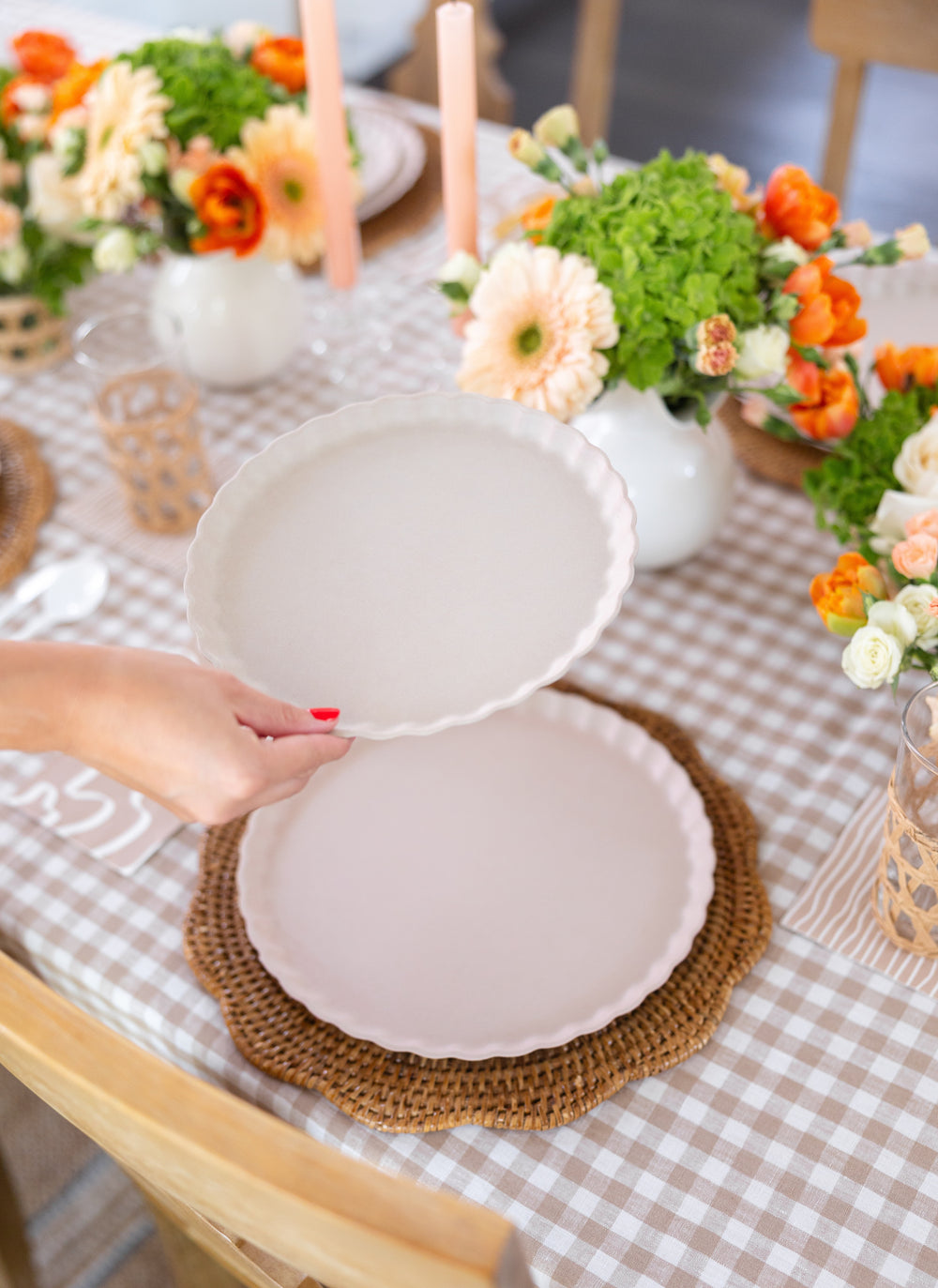 A hand with red nail polish sets down a Sophistiplate Simply Eco Compostable Table Setting in Sand onto a woven placemat on a gingham tablecloth, with floral centerpieces and candles in the background.