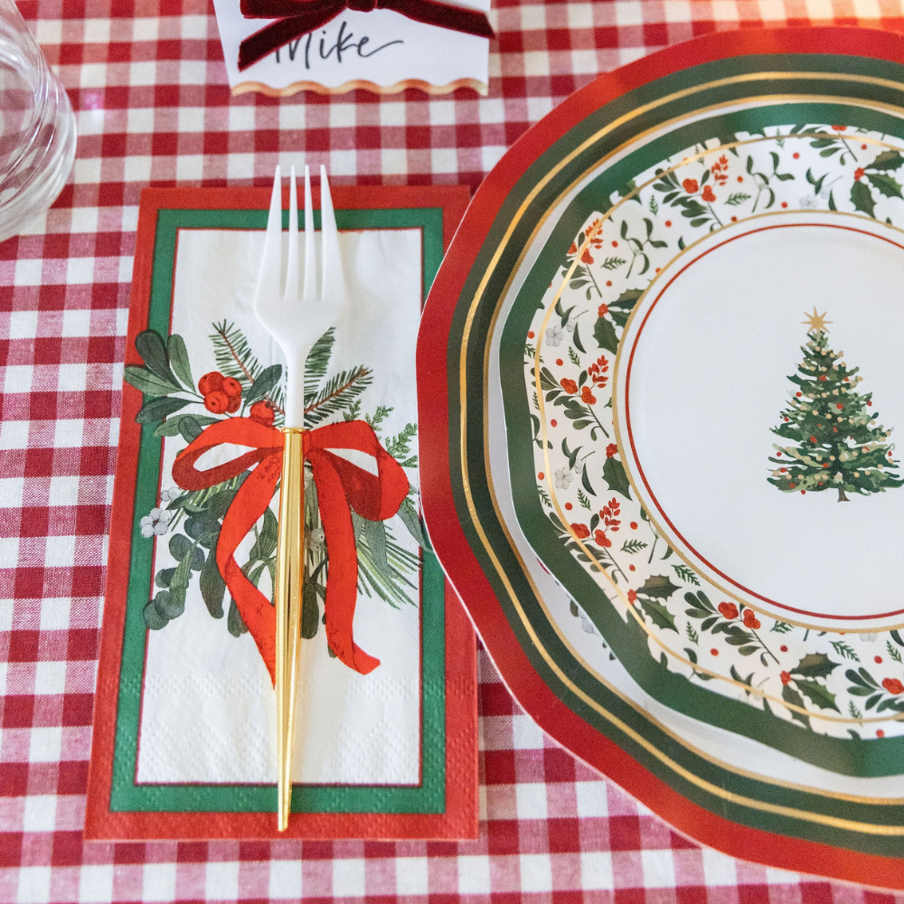 A Home for the Holidays: Christmas Table for 16 place setting on a red and white checkered tablecloth featuring a decorative paper plate with a Christmas tree, a festive wreath napkin, a gold-handled plastic fork, and a name card reading Mike.