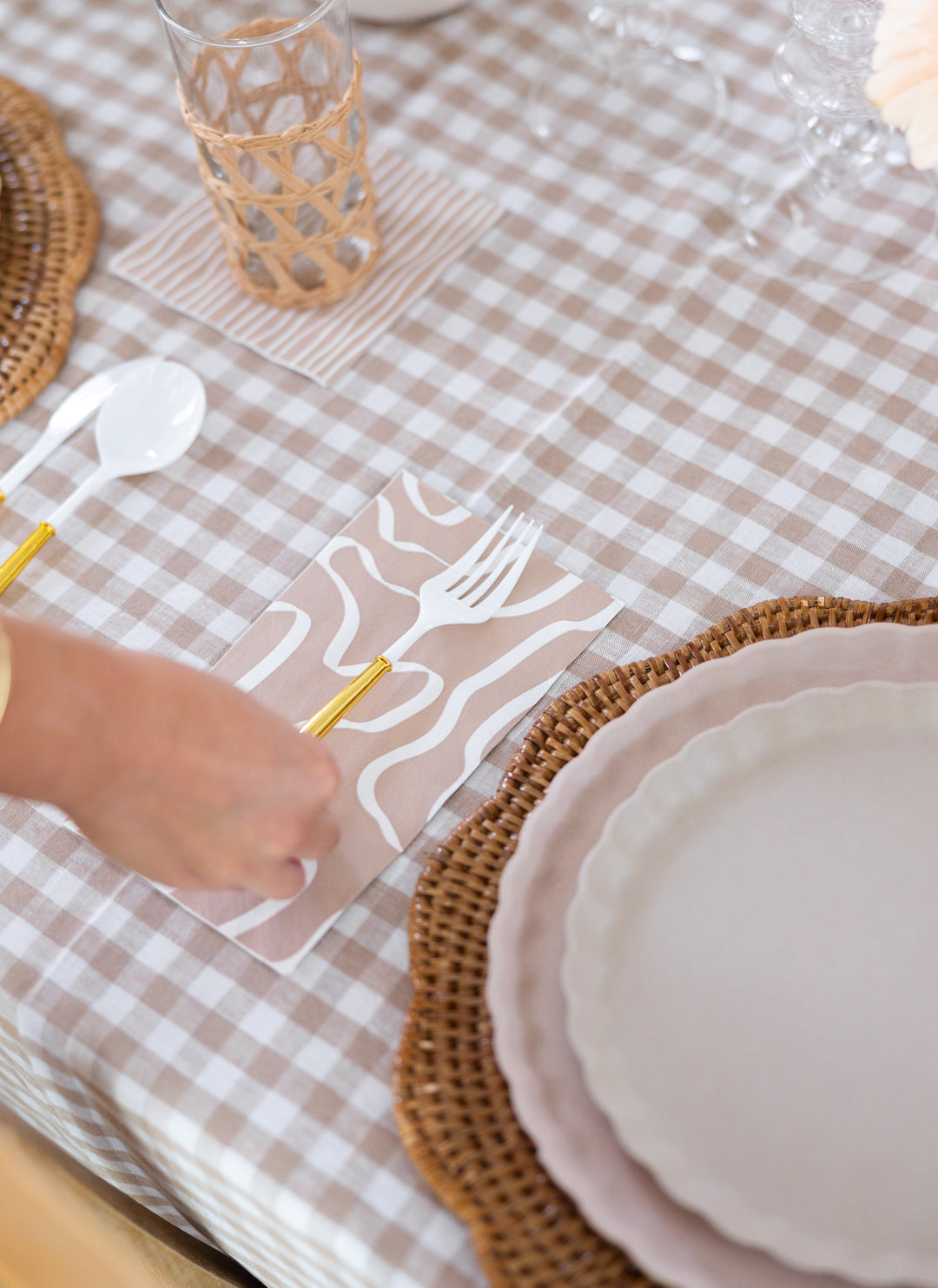 A close-up of a table set with a beige and white checkered tablecloth, woven placemat, scalloped plate, and someone placing Sophistiplate Guest Towel Eco Sand napkins (20/pkg) next to gold and white cutlery. A glass in a woven holder sits nearby.