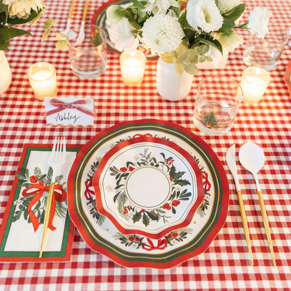A Home for the Holidays: Christmas Table for 16 setting on a red and white checkered tablecloth featuring festive paper plates with holly and red bows, an Ashley place card, gold-handled cutlery, and a small flower arrangement.