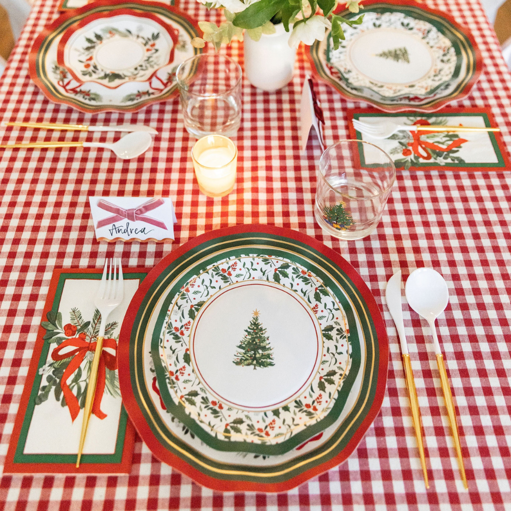 A Home for the Holidays: Christmas Table for 16 set on a red and white checkered tablecloth, featuring heavy duty paper plates, ribbon-print napkins, gold-handled cutlery, a glass, a small candle, and a name card labeled Andrea.