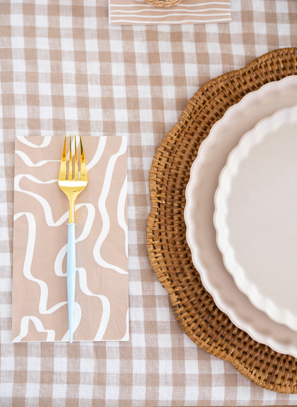 A setting with a beige and white checkered tablecloth, gold and blue fork on Sophistiplate Guest Towel Eco Sand napkins, and stacked white scalloped plates on a woven charger.