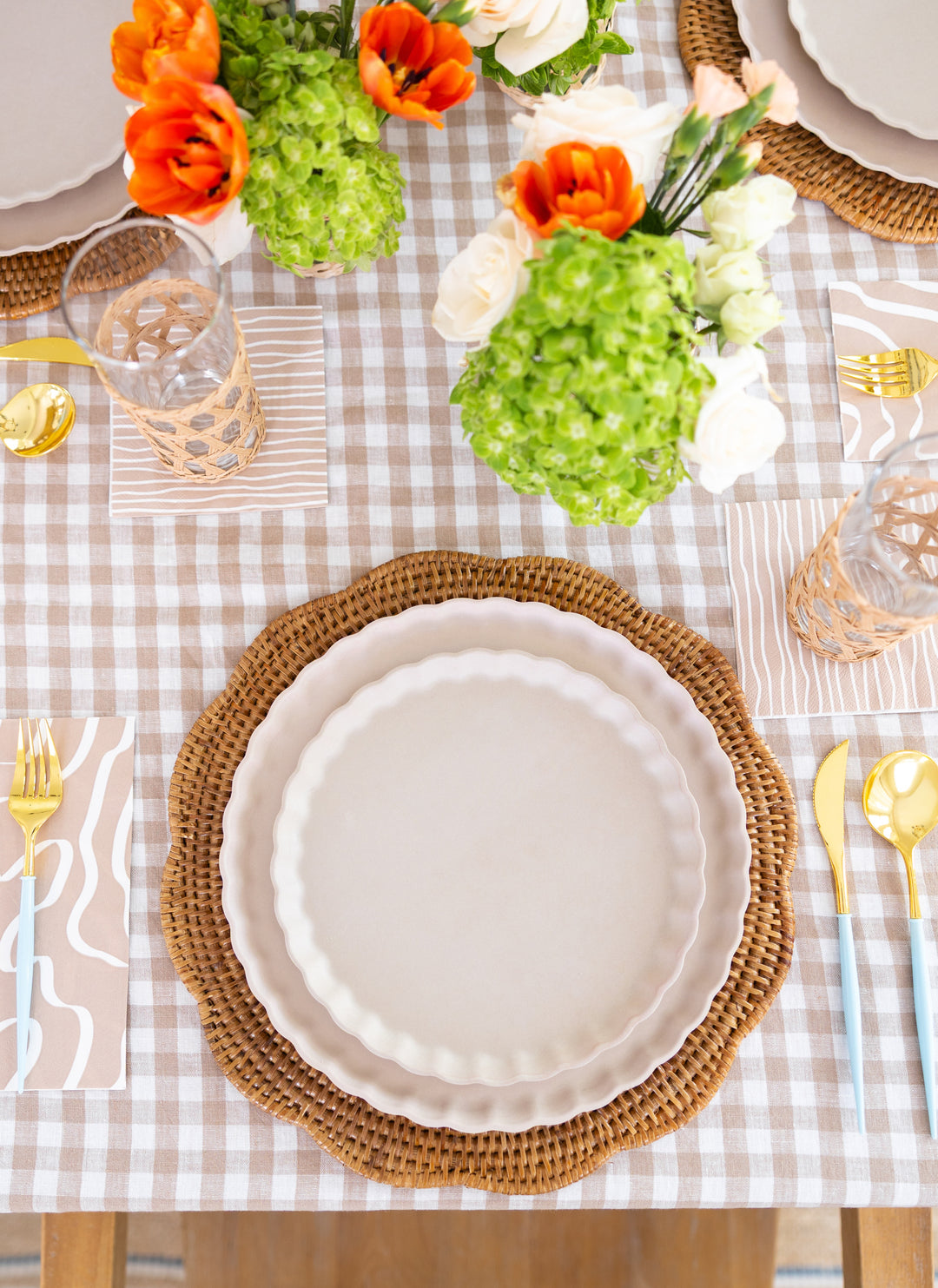 A table is set with white scalloped plates on woven placemats, patterned napkins, glasses, and Sophistiplate’s Bella Cutlery Gold/Sky Blue Handle. A light gingham tablecloth and vases of green and orange flowers complete the look.