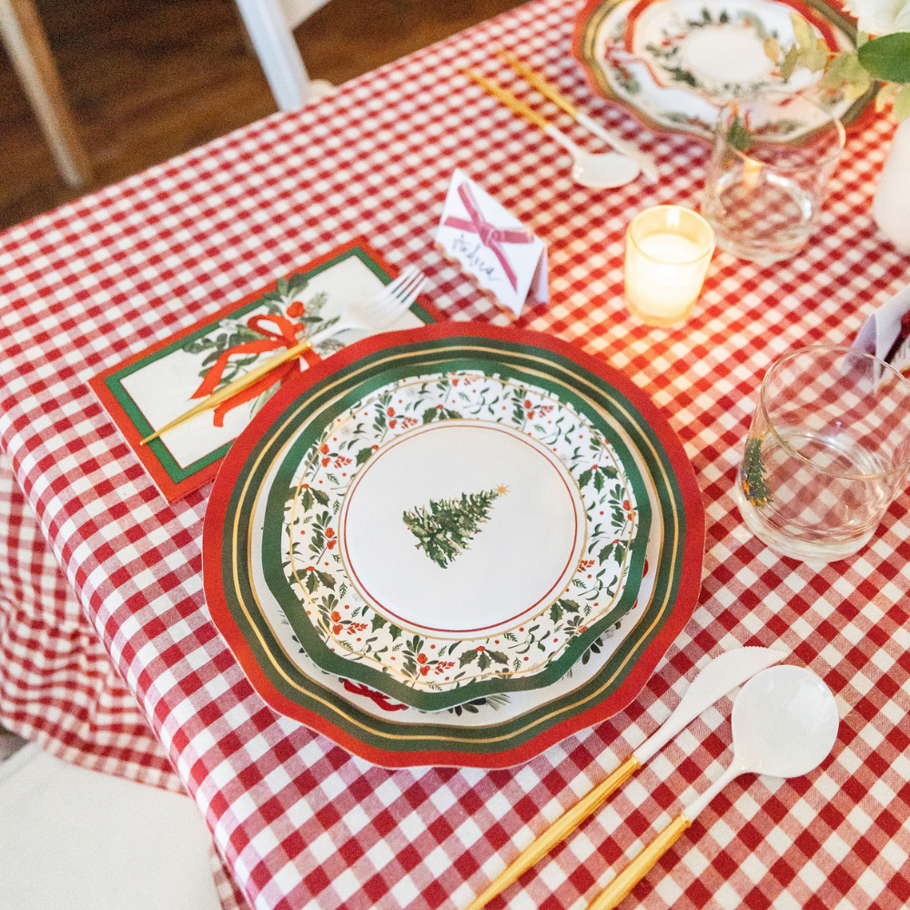 A Home for the Holidays: Christmas Table for 16 setting on a red and white checkered tablecloth, featuring layered Christmas tree paper plates, a holiday-themed napkin, a name card, a glass, and gold-handled cutlery.