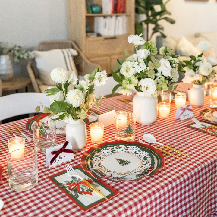 A Sophistiplate LLC Home for the Holidays: Christmas Table for 16 set with a red and white gingham tablecloth, decorated paper plates featuring a Christmas tree, floral centerpieces, lit candles, and place cards.