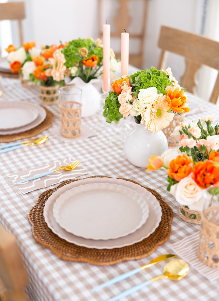 A spring-themed table setting with a gingham tablecloth, neutral plates on woven chargers, Sophistiplate Bella Cutlery Gold/Sky Blue Handle utensils, floral centerpieces with orange and white flowers, candles, and woven glasses.