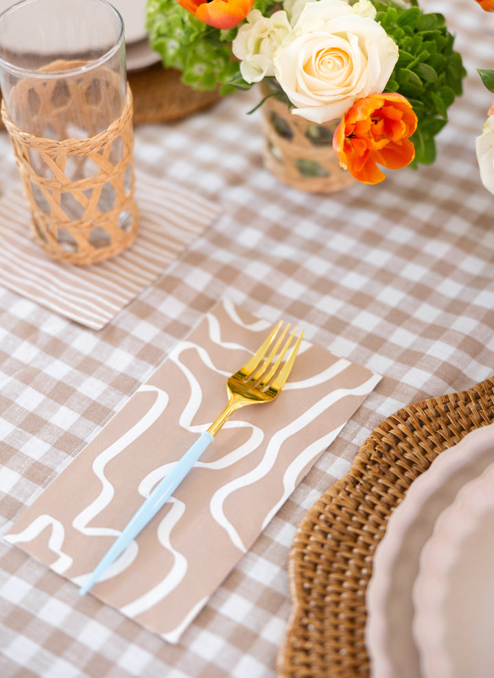 A gold fork with a light blue handle rests on Sophistiplate Guest Towel Eco Sand napkins atop a checkered tablecloth, next to a woven placemat, pink scalloped plate, glass with wicker holder, and vase of orange and white flowers.