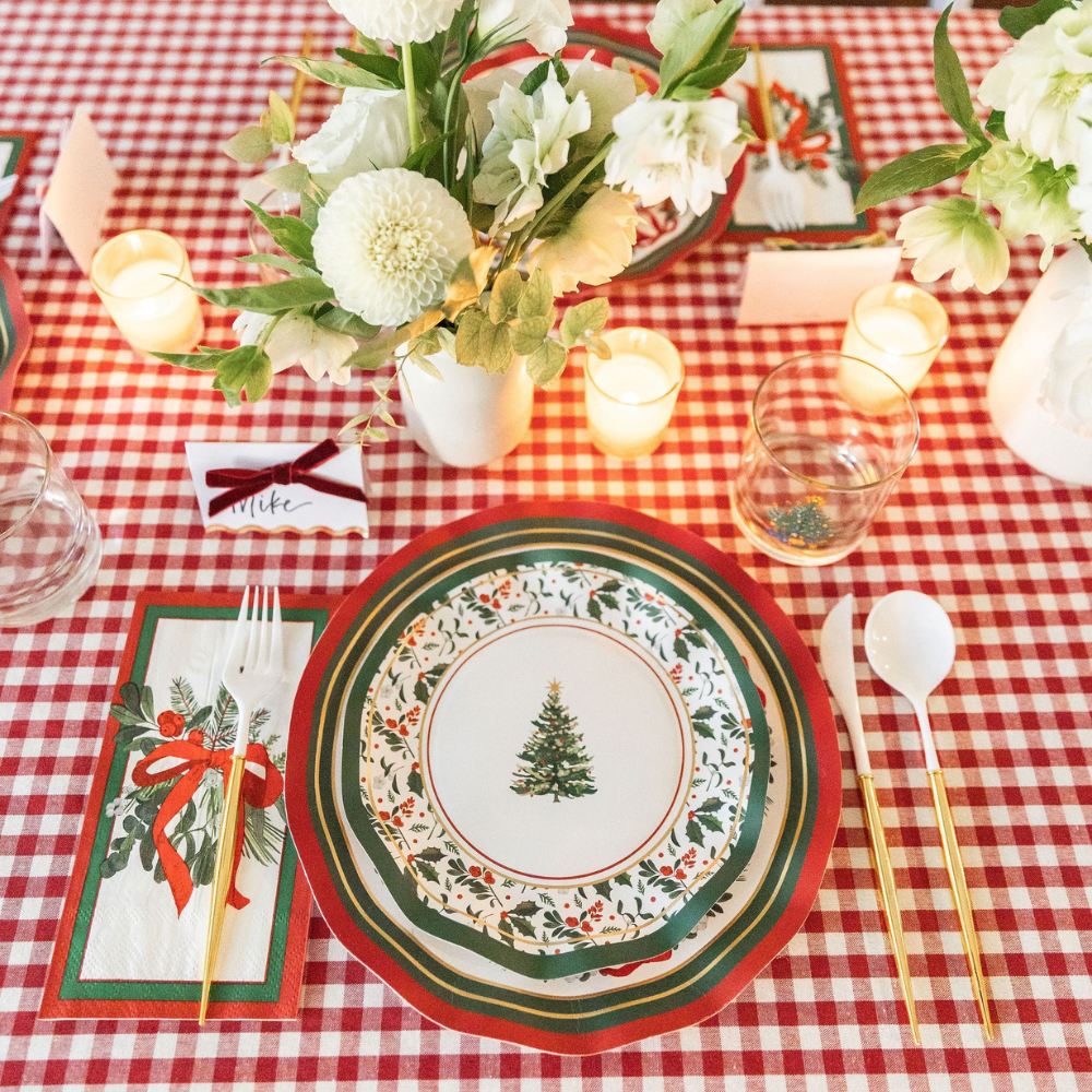 A Home for the Holidays: Christmas Table for 16 setting on a red and white checkered tablecloth, featuring Christmas tree paper plates, a floral napkin with a red bow, gold-trimmed cutlery, a name card, and a vase of white flowers.