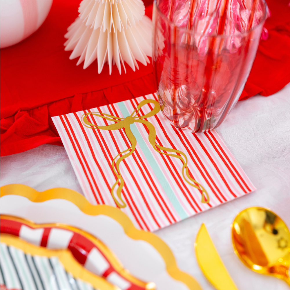 A square paper napkin with red and white stripes and a gold foil bow from the Tis the Season: Christmas Table for 16 collection by Sophistiplate LLC, set on a table with gold-rimmed designer paper plates and cutlery.
