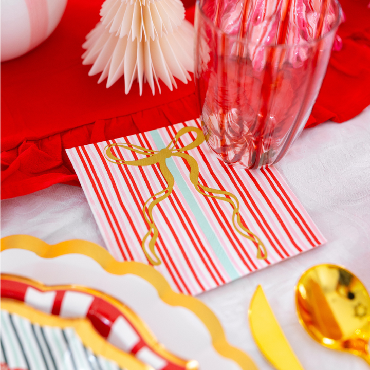 A square paper napkin with red and white stripes and a gold foil bow from the Tis the Season: Christmas Table for 16 collection by Sophistiplate LLC, set on a table with gold-rimmed designer paper plates and cutlery.