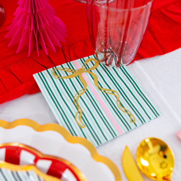Square paper napkin from the Tis the Season: Christmas Table for 16 set, featuring a green and white striped pattern and gold foil ribbon, arranged on a table with a red runner, white plate, and gold-toned cutlery.