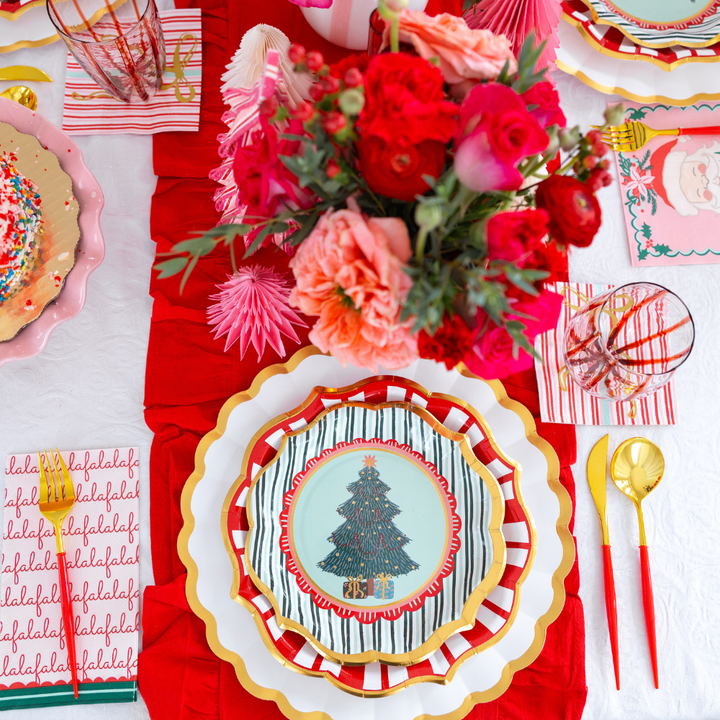 A Sophistiplate LLC Tis the Season: Christmas Table for 16 setting featuring designer paper plates with a tree illustration, layered atop striped and gold-rimmed plates, with red-handled gold cutlery, a patterned napkin, and a floral centerpiece.