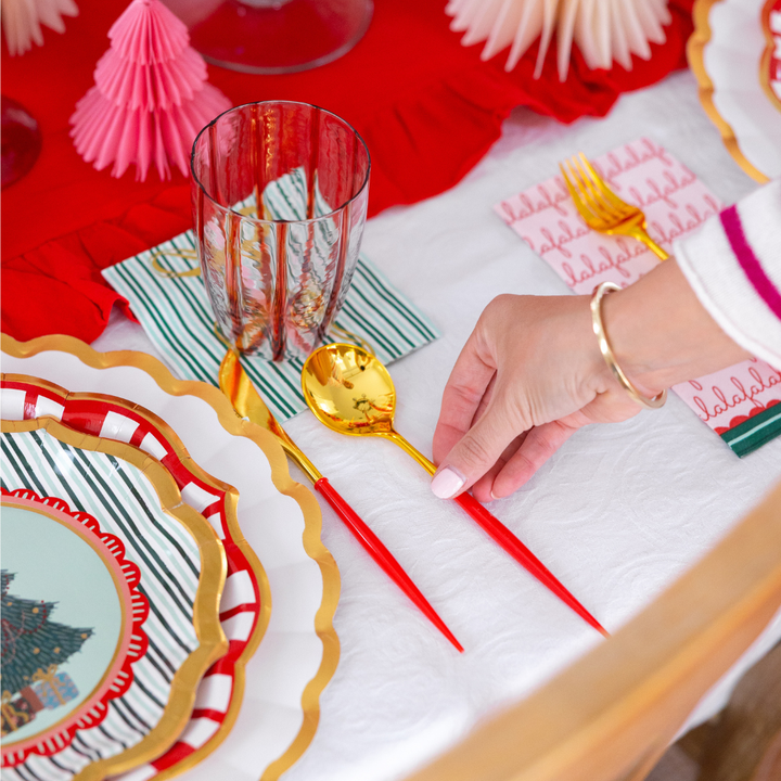 A hand places a gold-colored spoon with a red handle onto a Tis the Season: Christmas Table for 16 setting featuring layered designer paper plates, a glass, and a napkin.