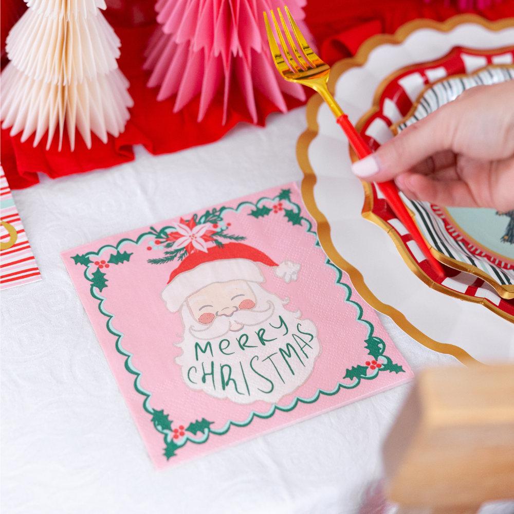 A pink Sugarplum Tis the Season Lunch Napkin/20pk featuring a smiling Santa Claus illustration and the text Merry Christmas sits on a table next to a scalloped plate and a gold fork.