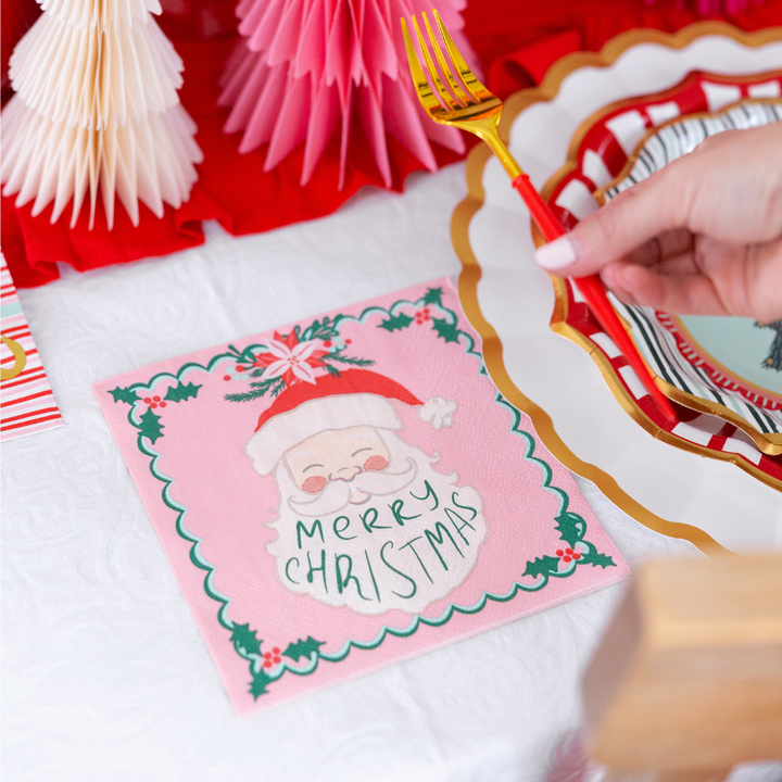 A pink Sugarplum Tis the Season Lunch Napkin/20pk featuring a smiling Santa Claus illustration and the text Merry Christmas sits on a table next to a scalloped plate and a gold fork.