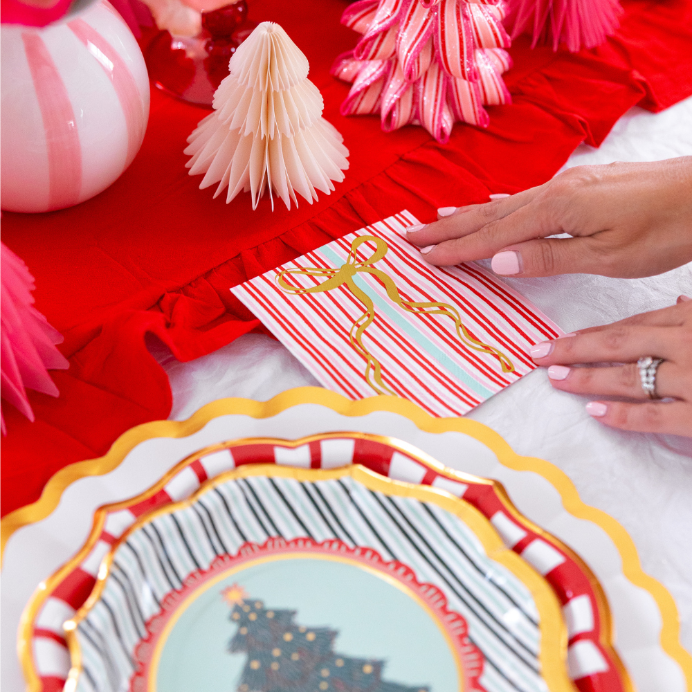 A person places a square card decorated with a gold ribbon onto a white table next to a stack of Tis the Season: Christmas Table for 16 designer paper plates and festive holiday ornaments.