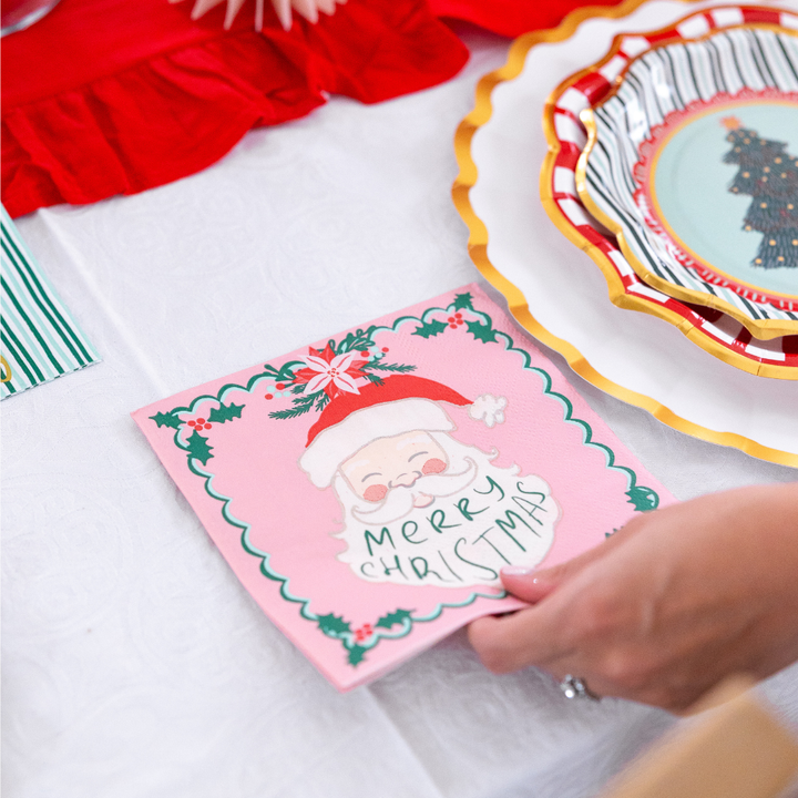 A hand places a square pink napkin with a Santa Claus illustration and the words Merry Christmas onto Sophistiplate LLC Tis the Season: Christmas Table for 16 paper plates at a holiday table setting.