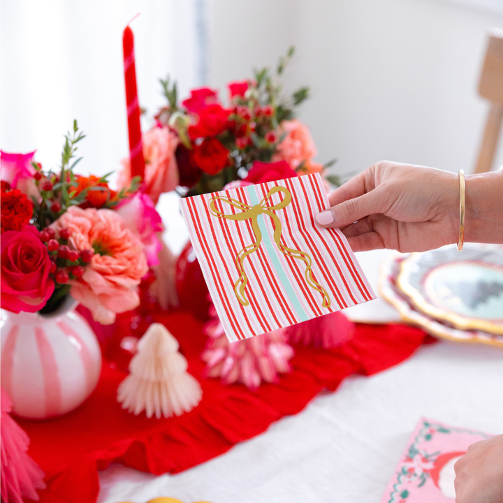 A hand holds a square, red and white striped Tis the Season: Christmas Table for 16 paper napkin with a gold ribbon design above a table decorated with flowers and holiday decor.