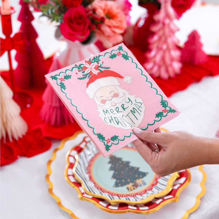 A hand holds a pink paper napkin from the Tis the Season: Christmas Table for 16 set, featuring a vintage-style Santa Claus illustration and the text Merry Christmas.