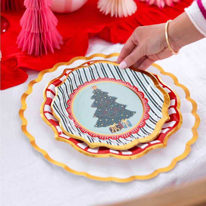 A hand places a small Christmas tree paper plate from the Tis the Season: Christmas Table for 16 set onto a stack of larger scalloped plates on a table set for the holidays.