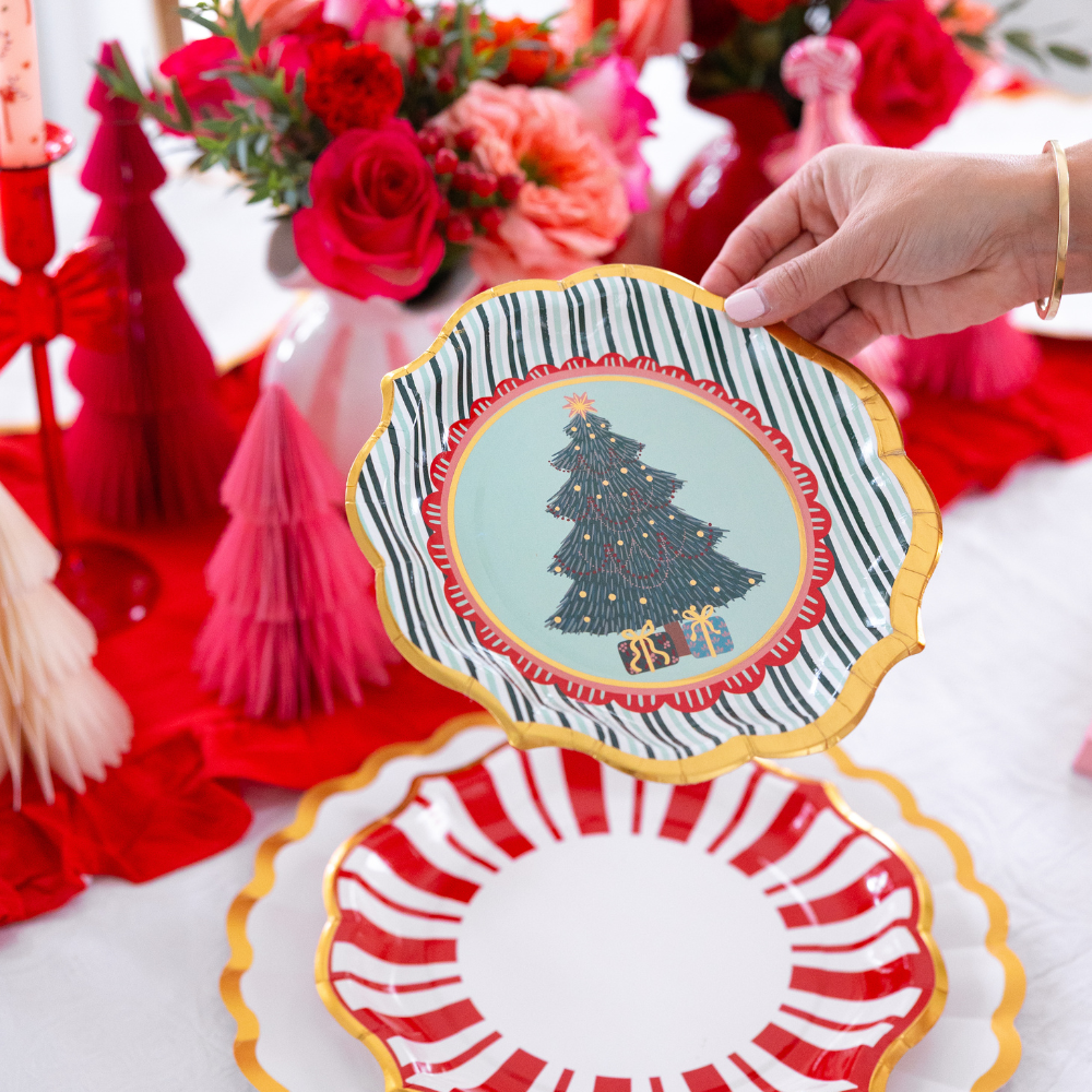 A hand holds a Christmas tree and green-striped plate from a Tis the Season: Christmas Table for 16 set above a red-striped plate, surrounded by floral arrangements and paper trees on a festive table.