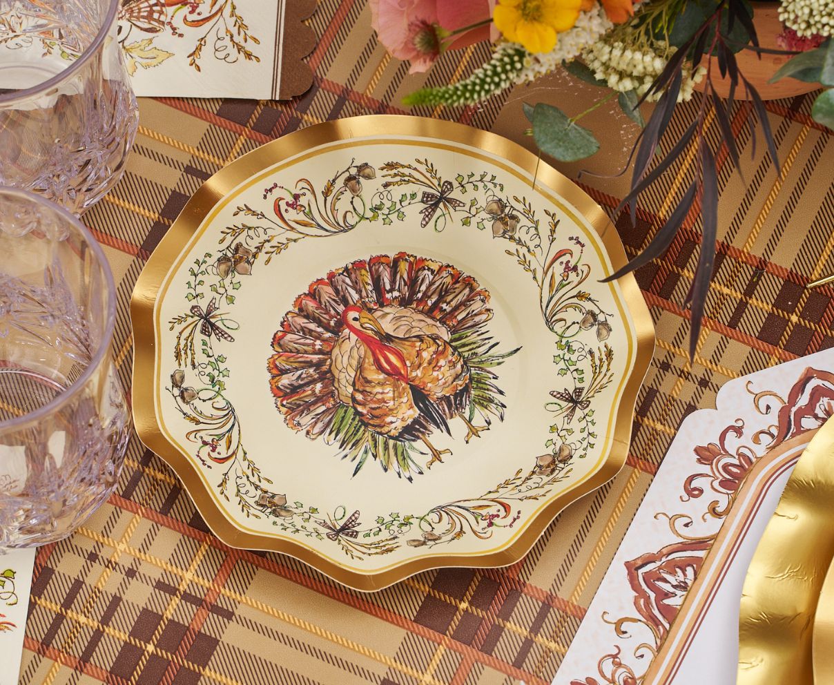 The Sophistiplate LLC Turkey & Gourds Table Setting Bundle features a decorative plate with a detailed turkey illustration at the center and a floral border, elegantly arranged on a plaid tablecloth alongside glassware and cutlery—a perfect example of designer disposable plates that blend festive style with convenience for any holiday gathering.