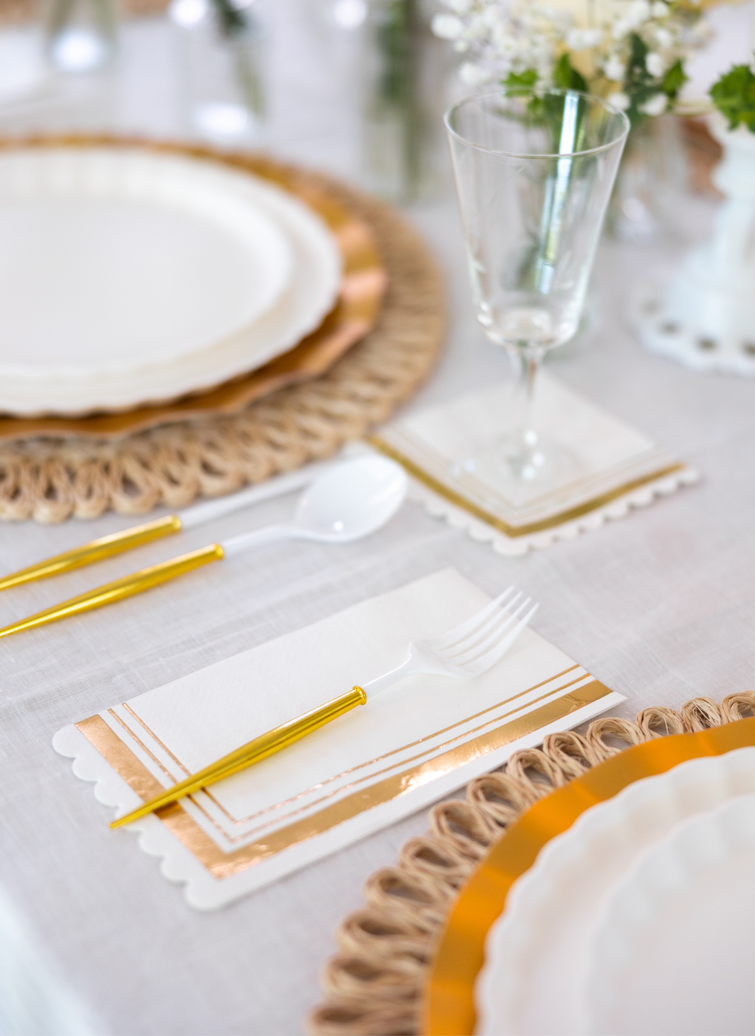 The Eco White & Gold Table Setting by Sophistiplate LLC features white designer disposable plates, gold and white cutlery, a glass, and a gold-accented napkin on a woven placemat atop a white tablecloth with floral centerpieces.