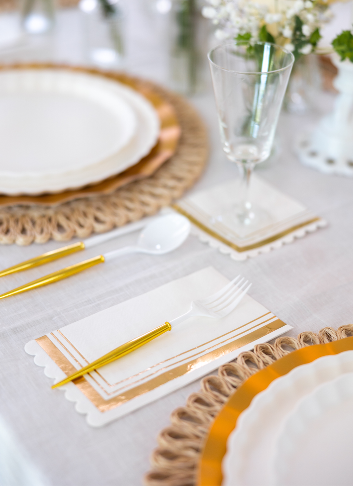 The Eco White & Gold Table Setting by Sophistiplate LLC features white designer disposable plates, gold and white cutlery, a glass, and a gold-accented napkin on a woven placemat atop a white tablecloth with floral centerpieces.