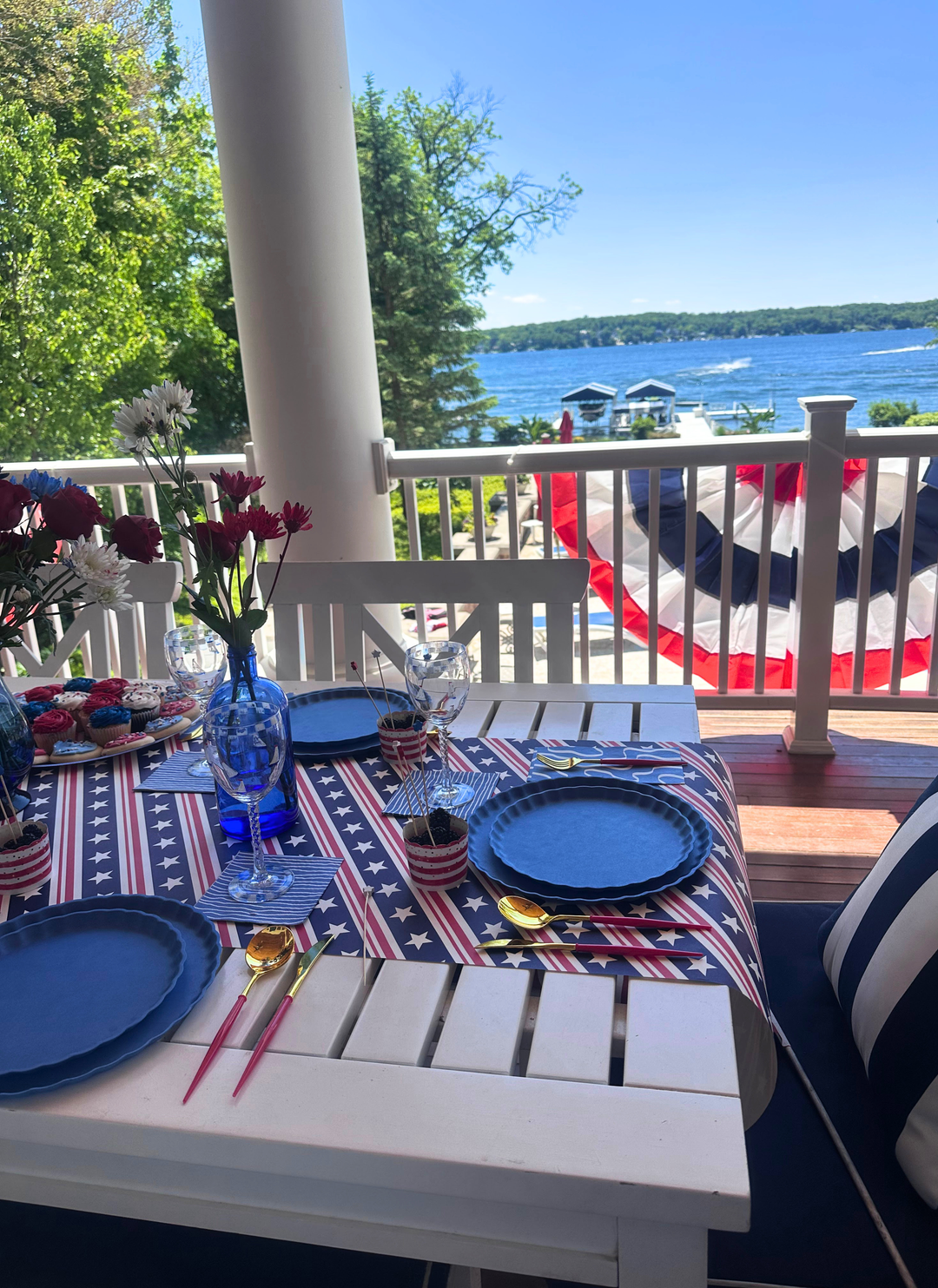 The Sophistiplate LLC 4th of July Table for 16+ in French Blue features eco-friendly plates, gold and red utensils, florals, drinks, and patriotic bunting—perfect for a festive celebration by the lake.
