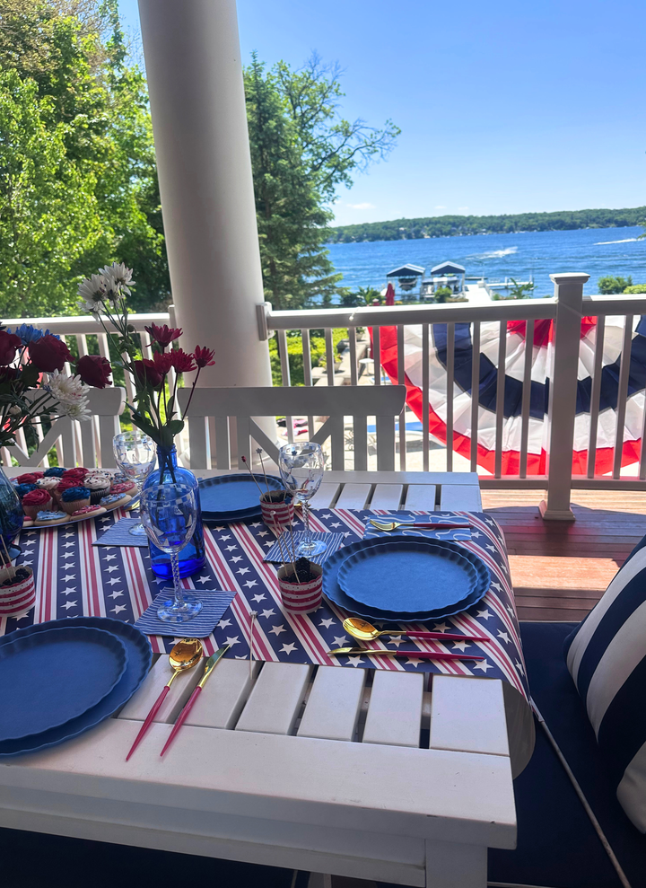 The Sophistiplate LLC 4th of July Table for 16+ in French Blue features eco-friendly plates, gold and red utensils, florals, drinks, and patriotic bunting—perfect for a festive celebration by the lake.